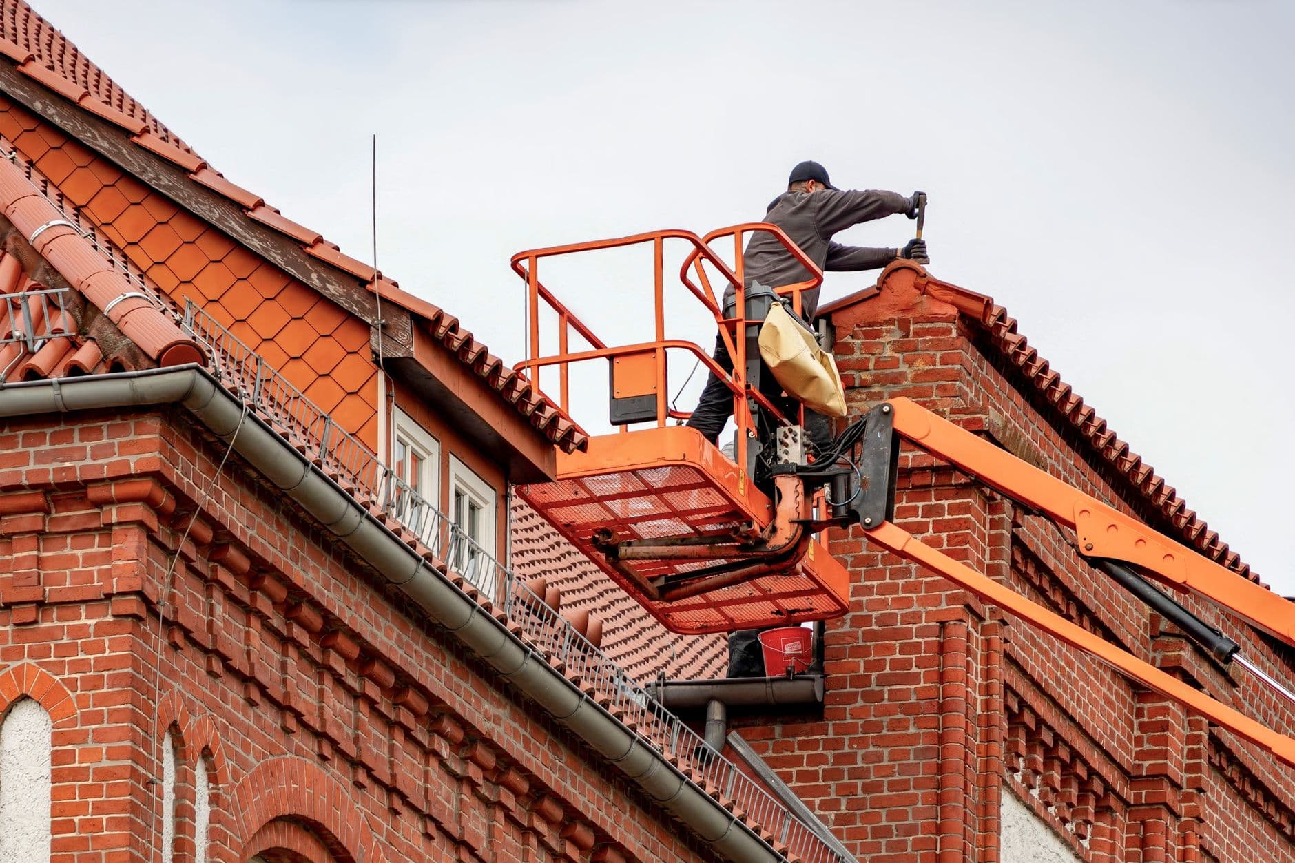 Dakwerker in een oranje hoogwerker aan het pannendak van een oud rood bakstenen gebouw
