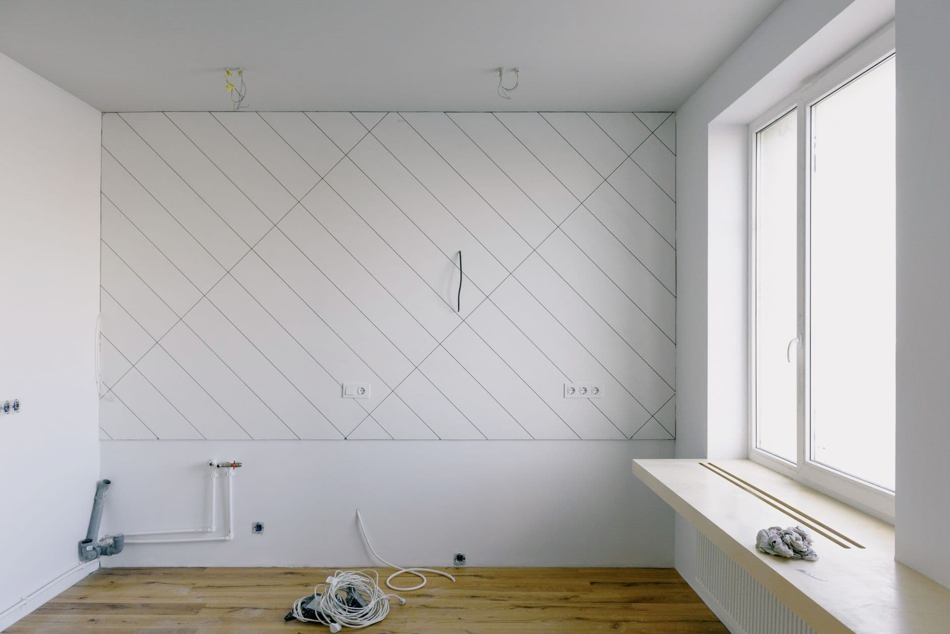 Lege witte ruimte tijdens de werken, met leidingen onderaan de muur en diagonale lijnen op de wand