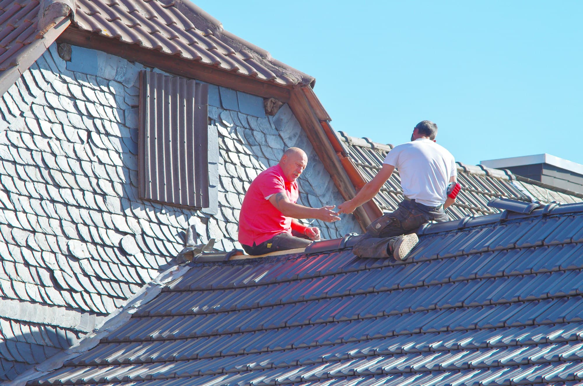 Twee mannen aan het werk op een dak met blauwgrijze dakpannen, naast een met leien beklede muur
